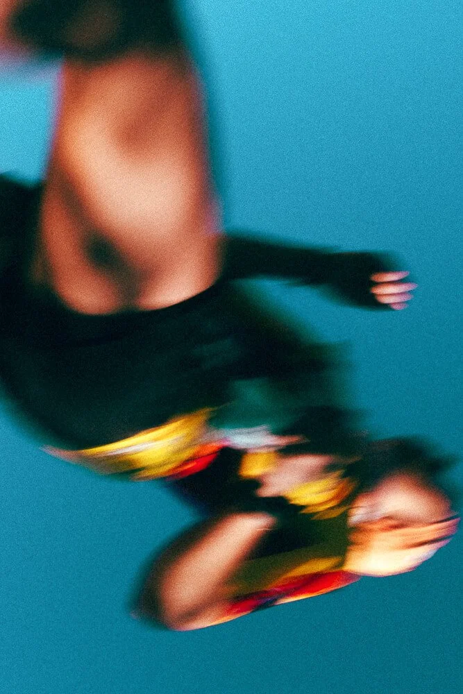 A person performing a dance move with their body bent back, over a reflective surface, wearing a black top and colorful pants, against a blue background.