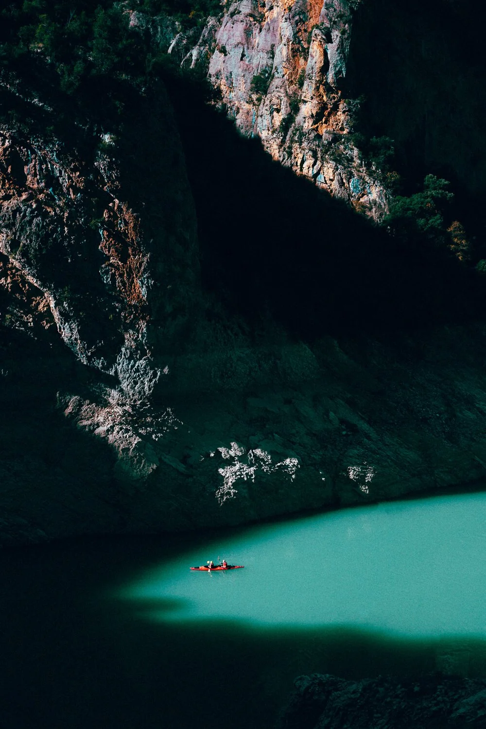Une petite embarcation rouge avec deux personnes naviguant sur un lac vert entouré de hautes falaises rocheuses avec quelques arbres.