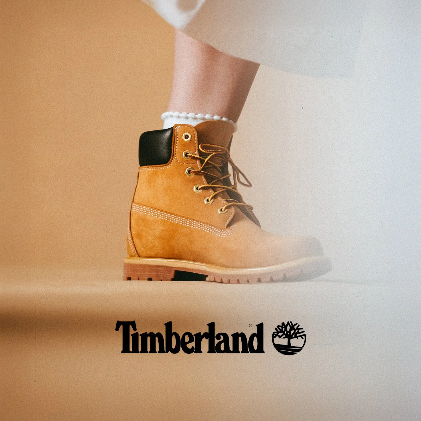 Une botte Timberland beige avec un renfort noir sur la cheville, portée avec une chaussette blanche à froufrous, vue de côté sur fond blanc et beige.