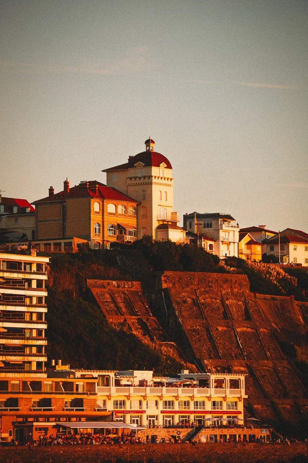Houses and buildings on a hill during sunset, with a prominent tower on top and a clear sky.