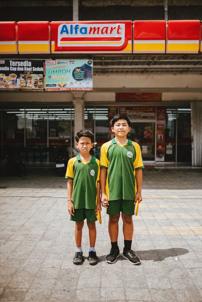 Two boys in green and yellow school uniforms standing outside an Alfamart store.