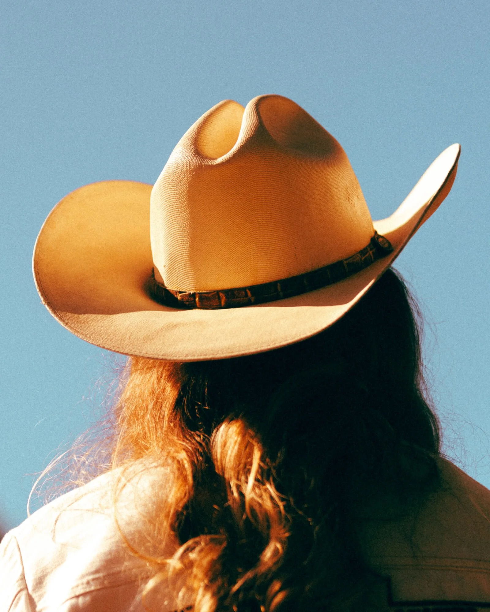 Front view of a person wearing a beige cowboy hat with a dark band, standing outdoors against a clear blue sky, with long, wavy hair.