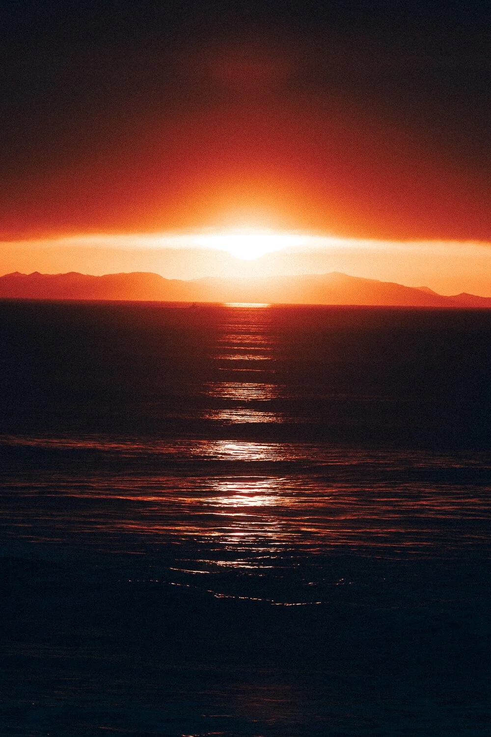 Sunset over the ocean with mountains in the background and clouds above, reflecting light on the water.
