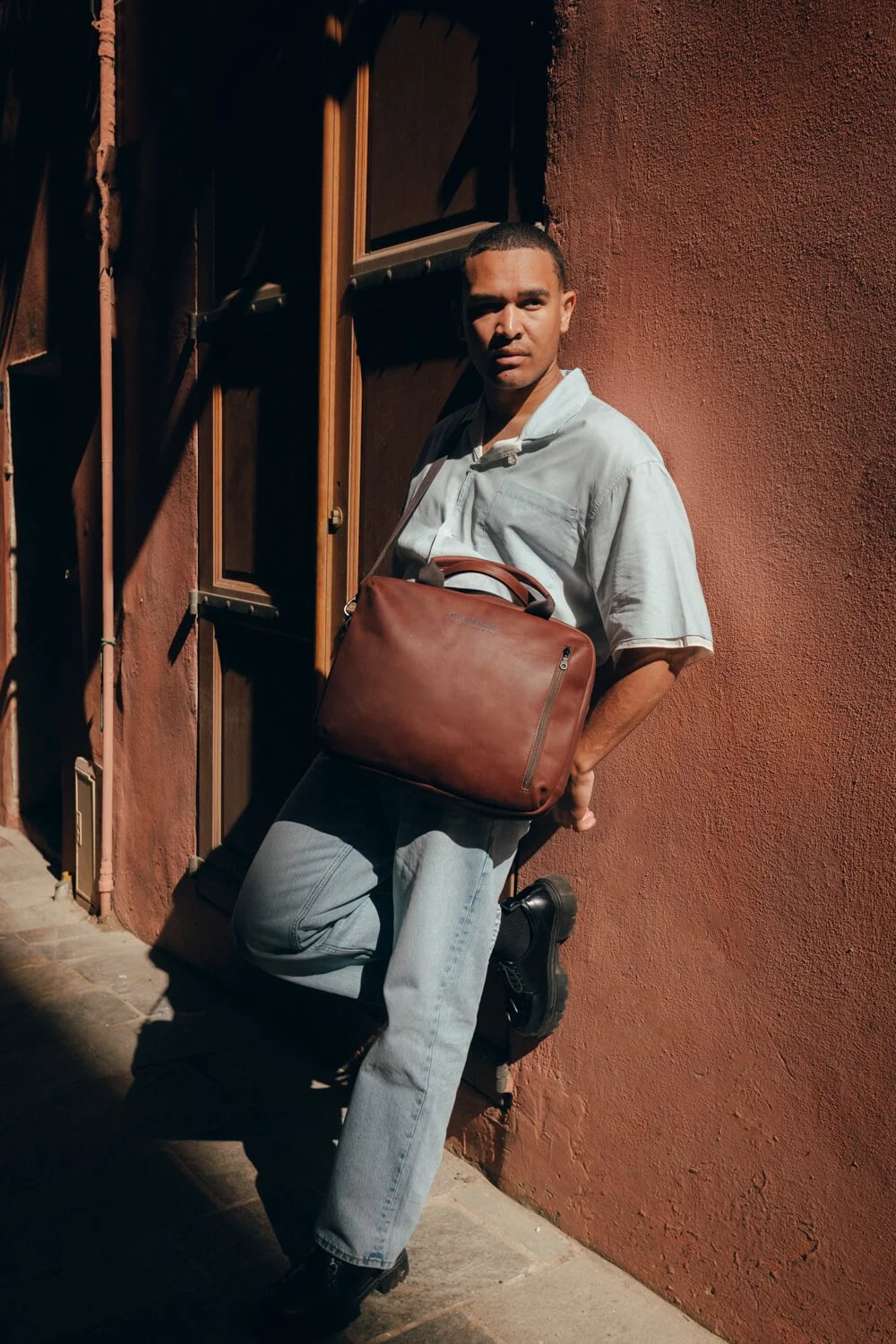 A man with short hair, wearing a white shirt and light blue jeans, standing against a red wall with his left knee bent and his right foot flat on the ground. He is holding a brown leather bag and looking to the side, with sunlight casting shadows on 