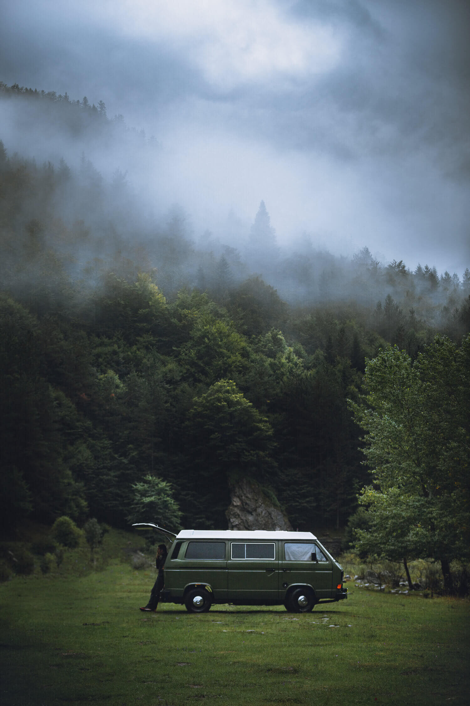 Une camionnette verte garée sur une pelouse devant une forêt brumeuse.