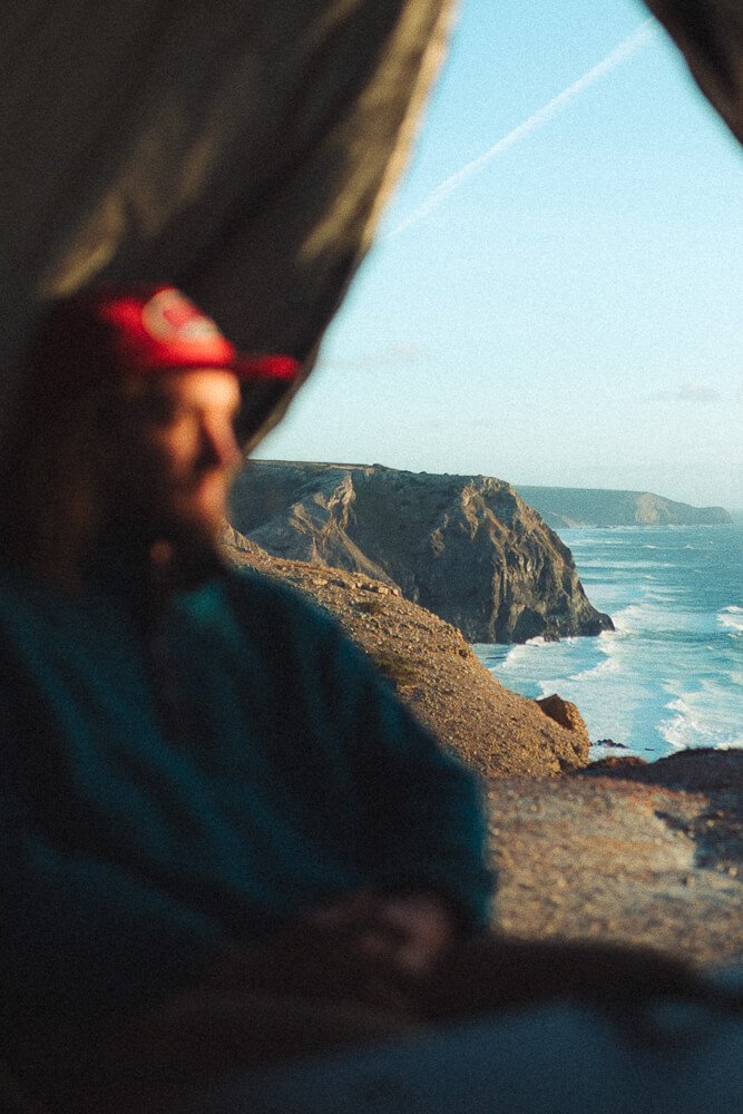 A person in a red hat sitting inside a tent, overlooking cliffs and the ocean during sunset.