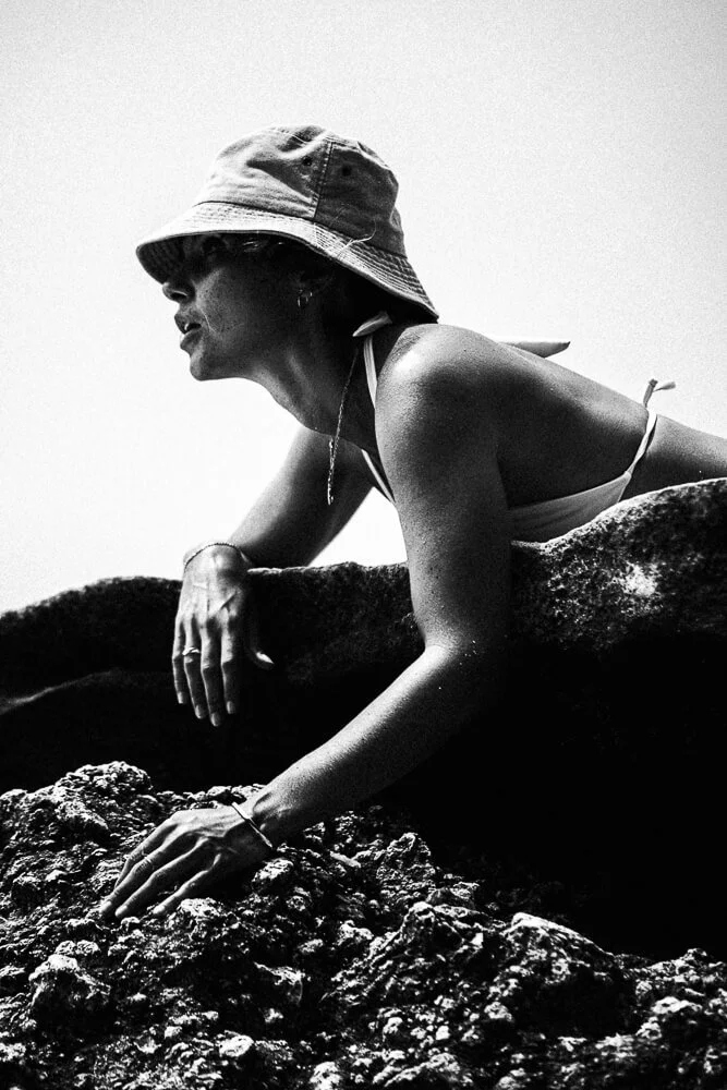 A woman wearing a wide-brimmed hat and a swimsuit leans on a rock formation, touching the rocks with her hand, outdoors.