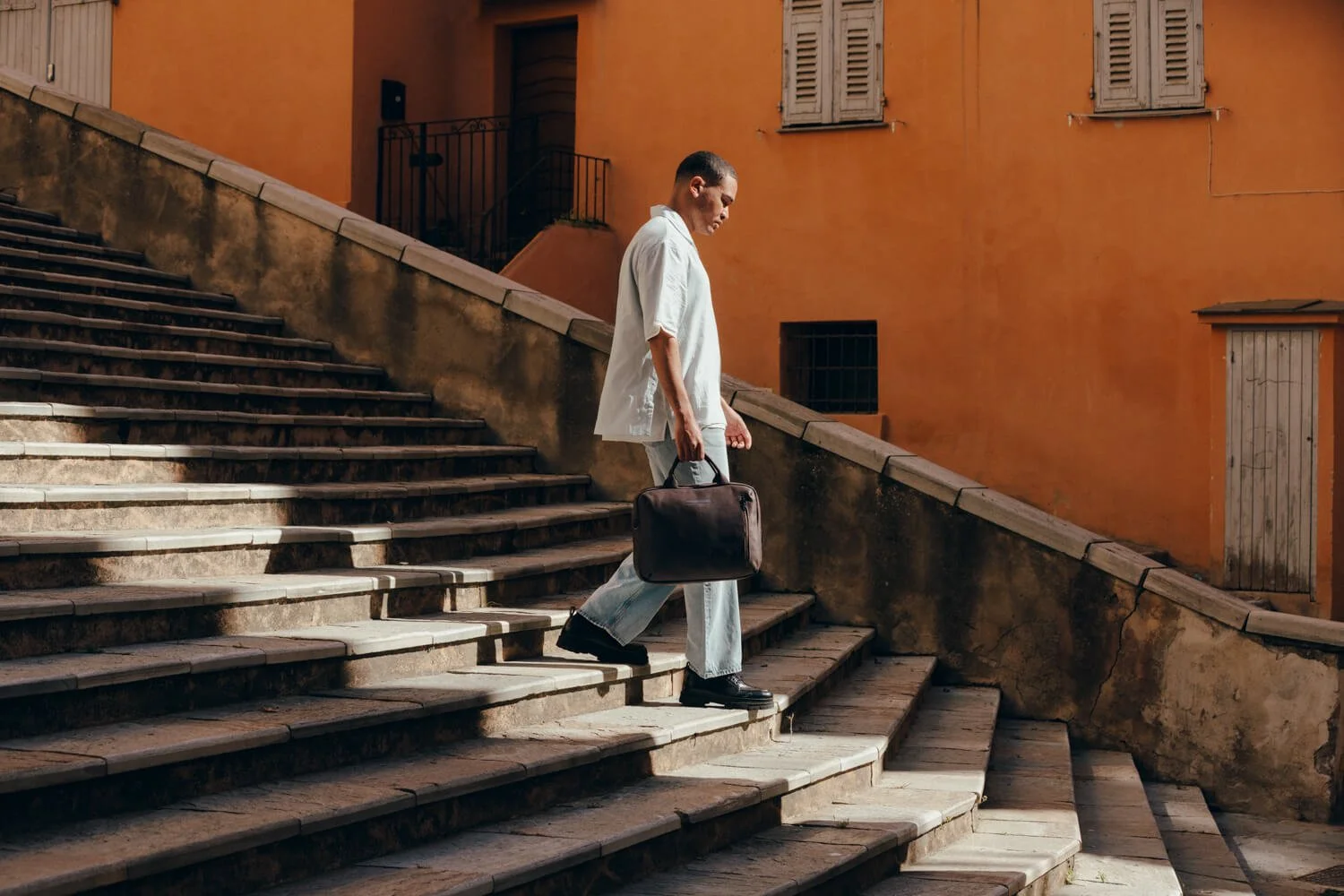 A man walking down a stone staircase with a black briefcase in hand, against an orange building background.