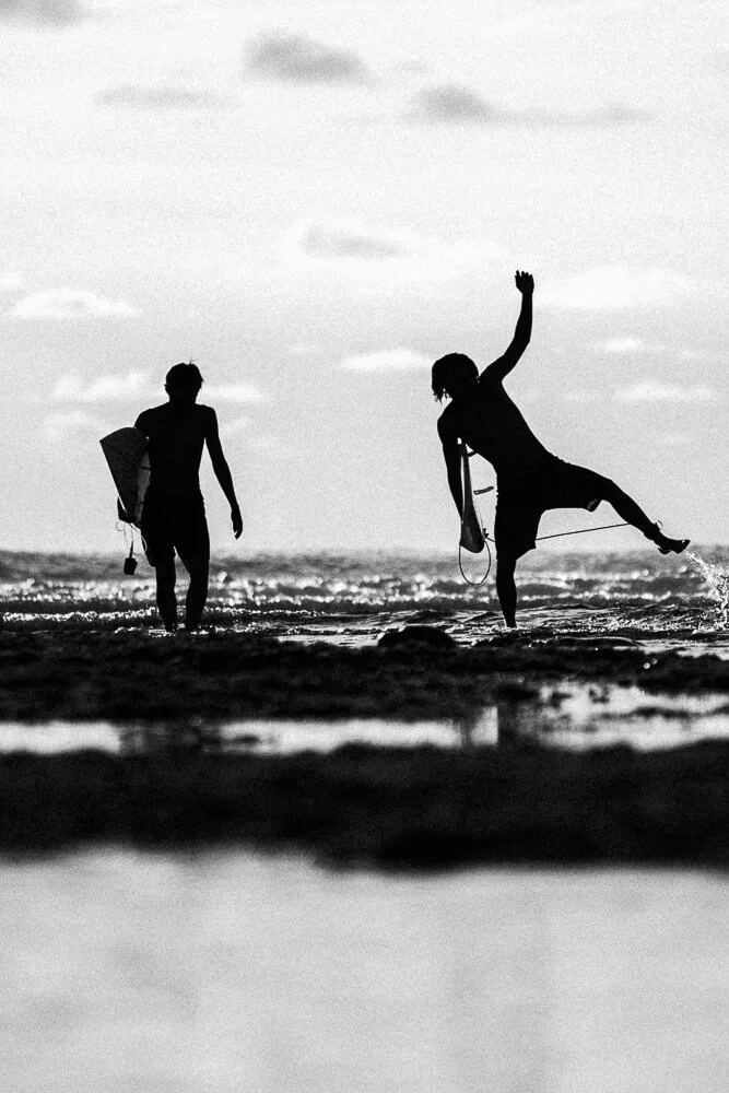 Silhouettes of two people carrying surfboards walking on the beach with the ocean in the background, one person doing a surfing pose.