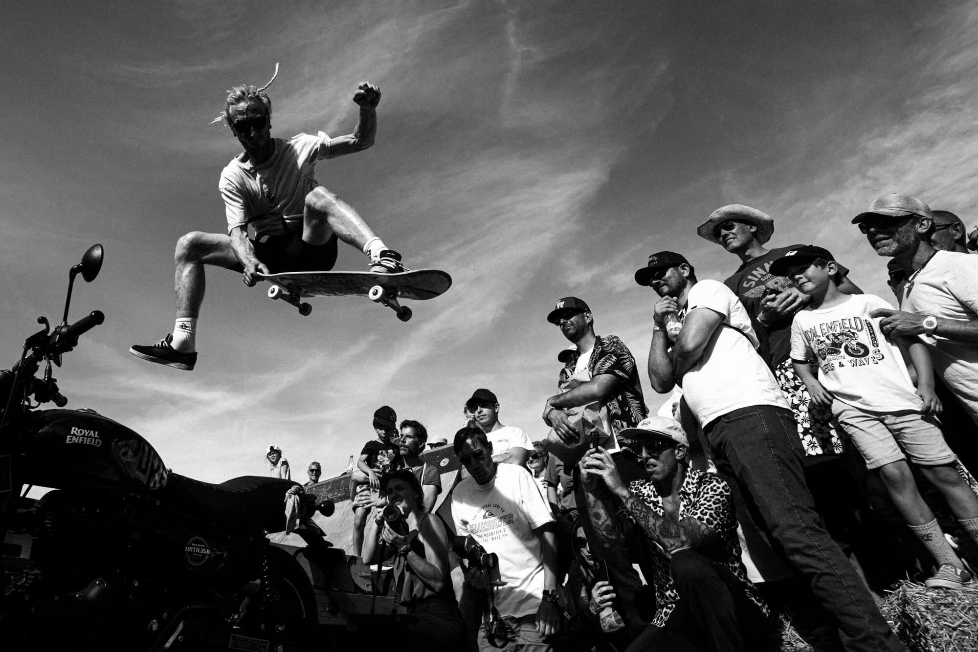 A person performing a skateboard trick in mid-air, surrounded by a crowd of onlookers watching at an outdoor event on a sunny day.