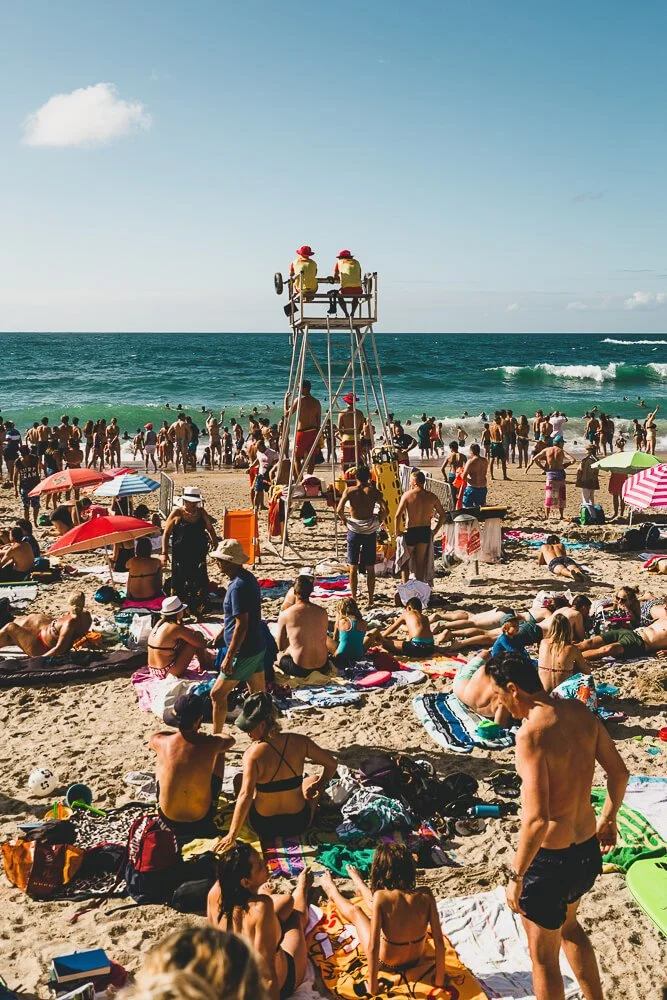 Crowded beach scene with many people sunbathing, sitting under umbrellas, and enjoying the water. Lifeguards in yellow shirts are on a tall chair, watching over the beach. The ocean has small waves, and the sky is clear with a few clouds.