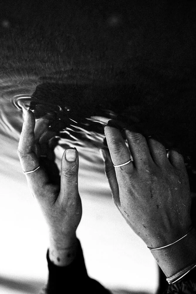 Two hands with rings touch the surface of a liquid, creating ripples in the water.