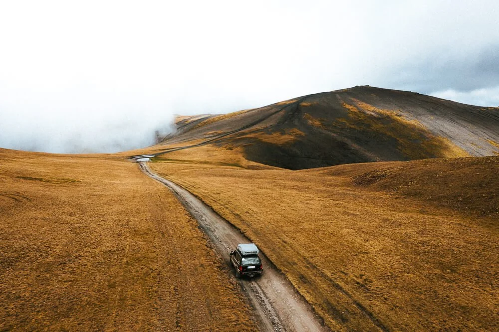 A black SUV driving on a narrow dirt road through an open, grassy landscape with rolling hills and mountains in the background under cloudy skies.