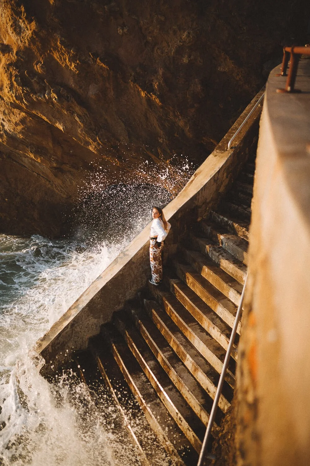 Woman standing on wet stairs beside the ocean at a rocky shoreline, with waves crashing nearby and a wet rock wall in the background.