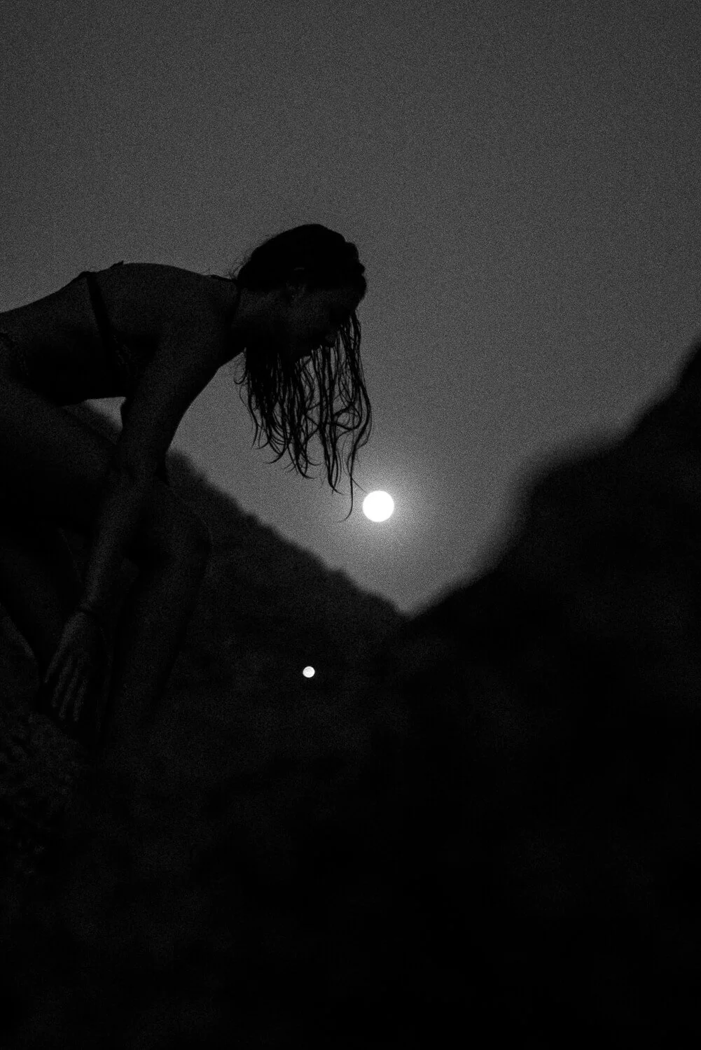 A person with wavy hair crouching down at night, with the moon visible in the sky and a second smaller moon or bright object nearby, silhouetted against the night sky.