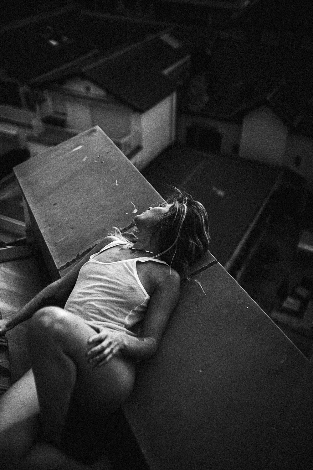 A woman in a white tank top lying on her side on a table in a dark room, with her eyes closed and head tilted back.