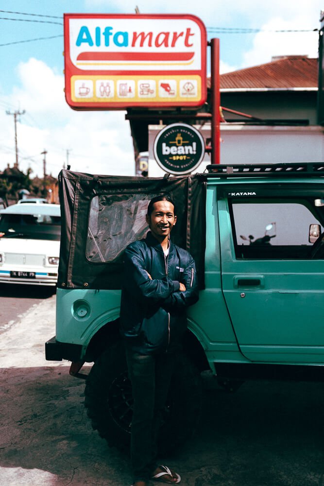 A man standing in front of a teal off-road vehicle with a black canopy, smiling and crossing his arms. Behind him is a sign for Alfa Mart, a convenience store, and a smaller sign for Bean Spot. There are other vehicles parked nearby.