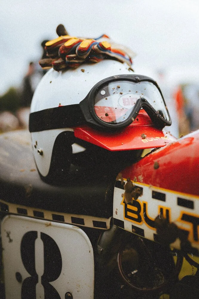 A motorcycle helmet with a racing visor, black goggles, and a multicolored scarf or cloth on top, resting on a racing bike with a red and white fairing and a visible number 8 plate, at an outdoor racing event.