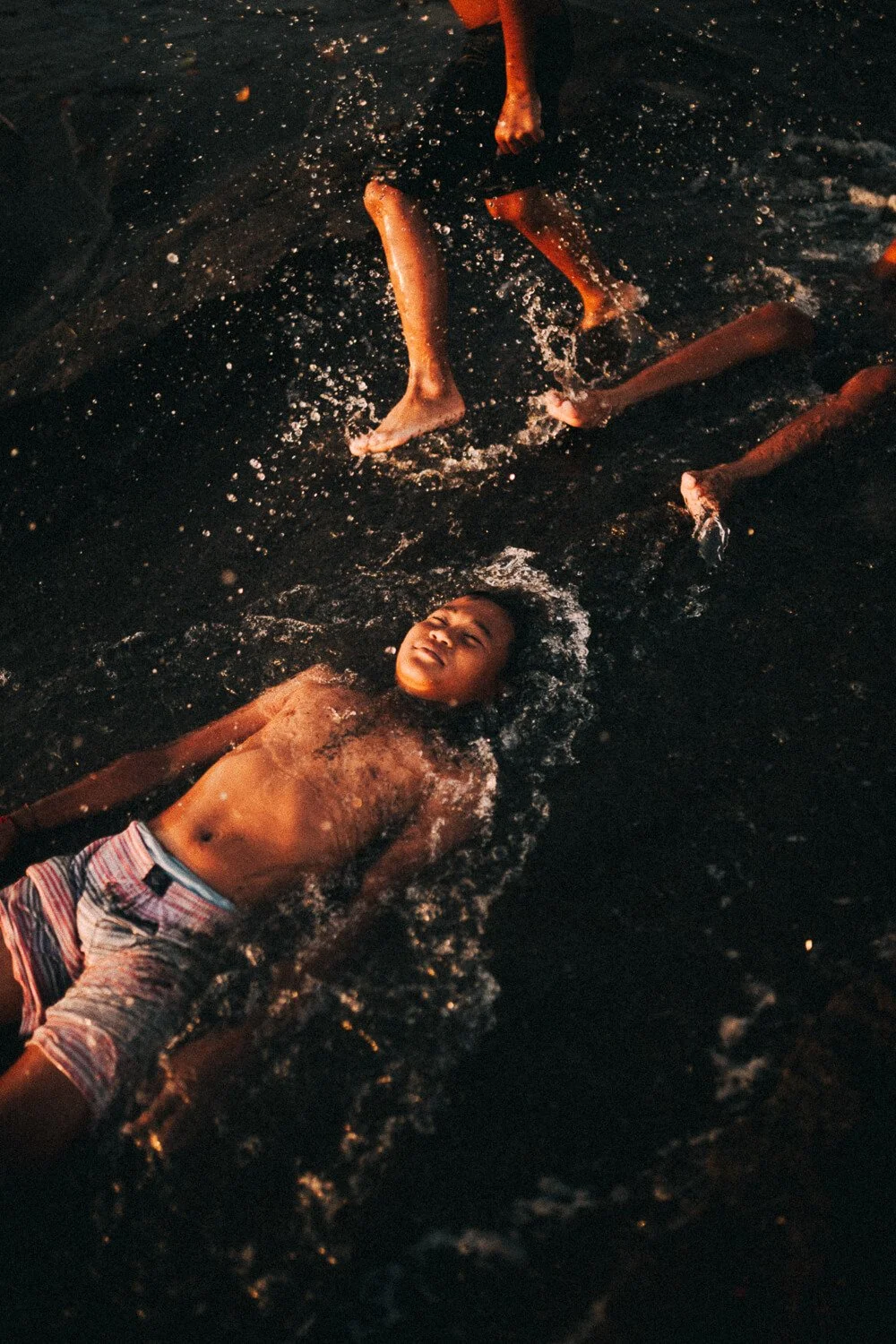 Un garçon allongé dans de l'eau, entouré de plusieurs autres personnes qui nagent ou jouent dans l'eau.