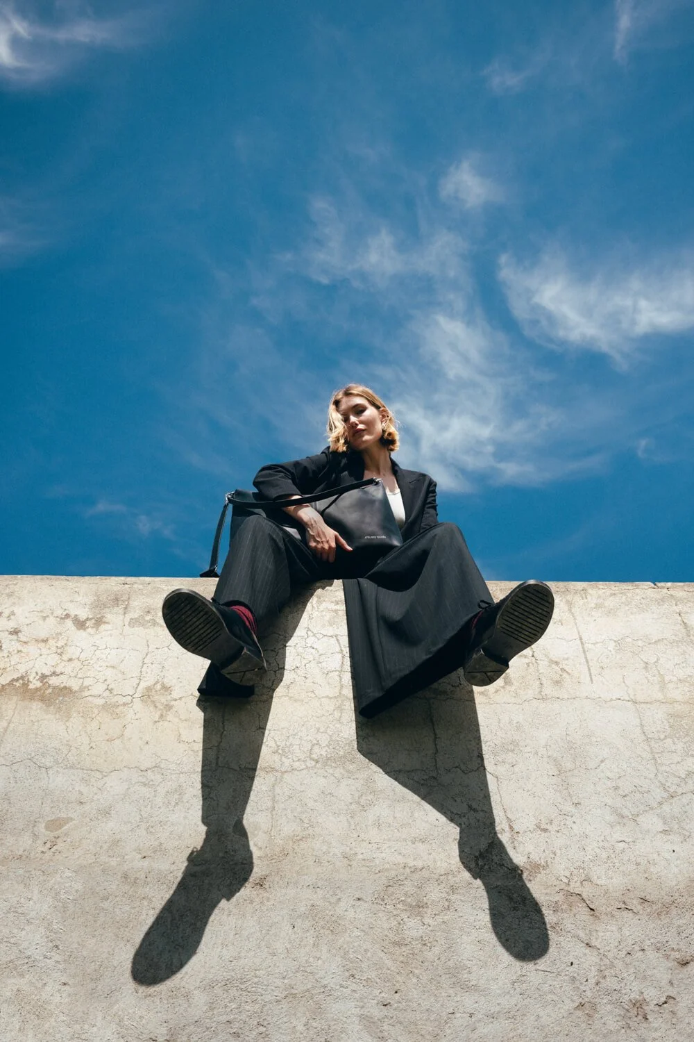 A woman in a black suit sitting on a concrete ledge with her feet hanging down, casting shadows on the wall, against a bright blue sky with wispy clouds.