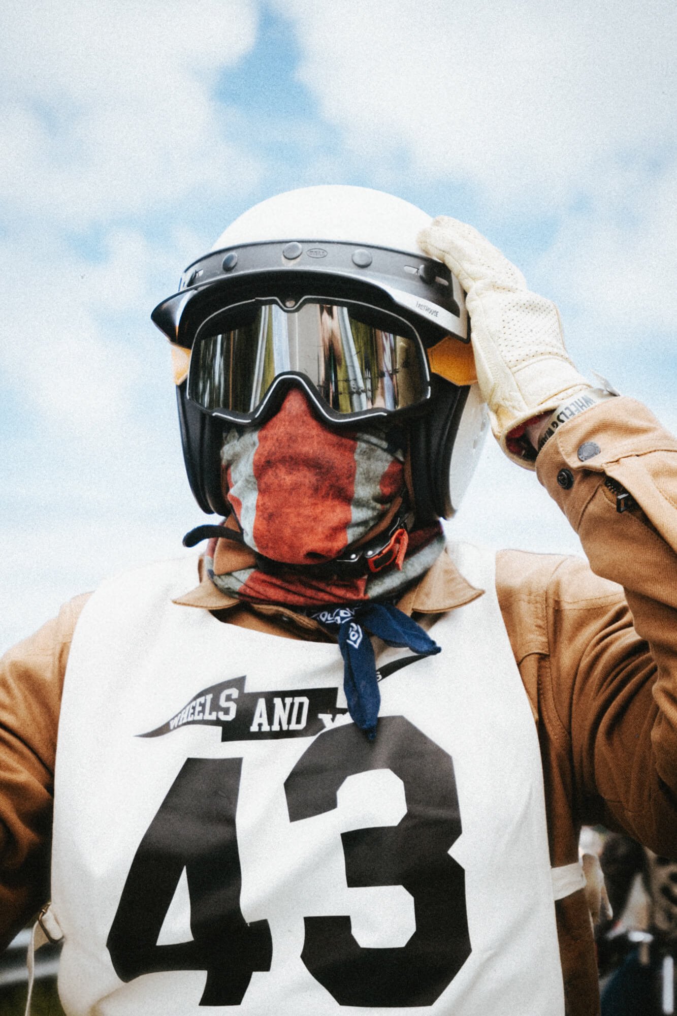 Person wearing a motocross helmet with goggles and a red neck gaiter, adjusting the helmet's strap, dressed in a brown jacket and a white vest with bold black lettering.