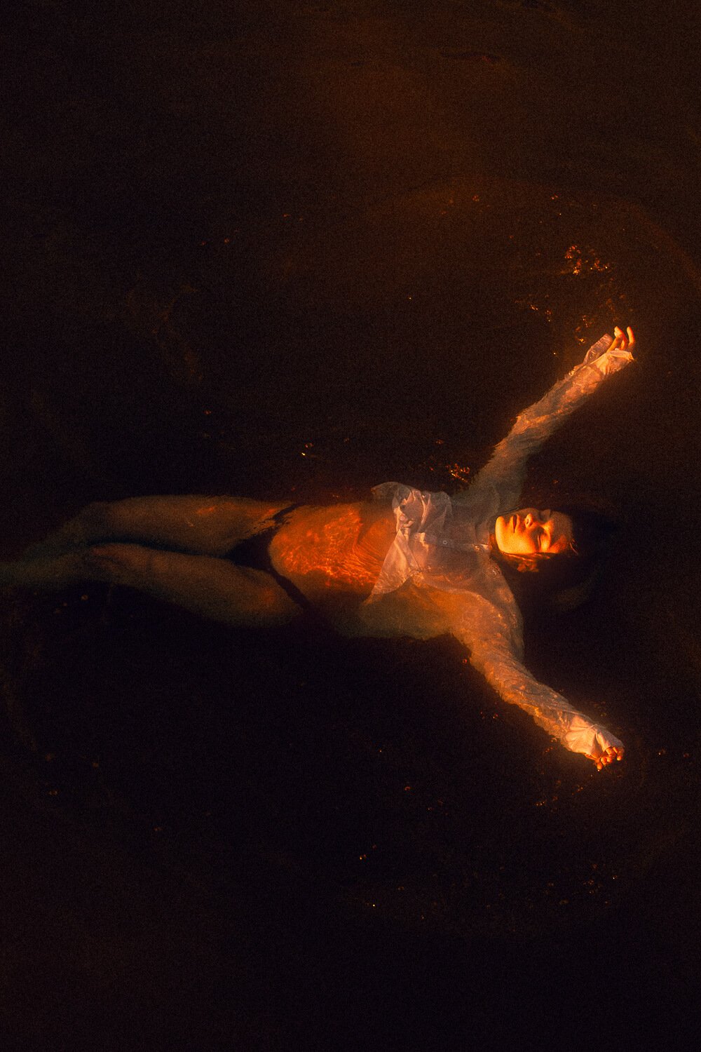 A woman lying in shallow water at night, illuminated by a warm light, with her eyes closed and arms extended outward.