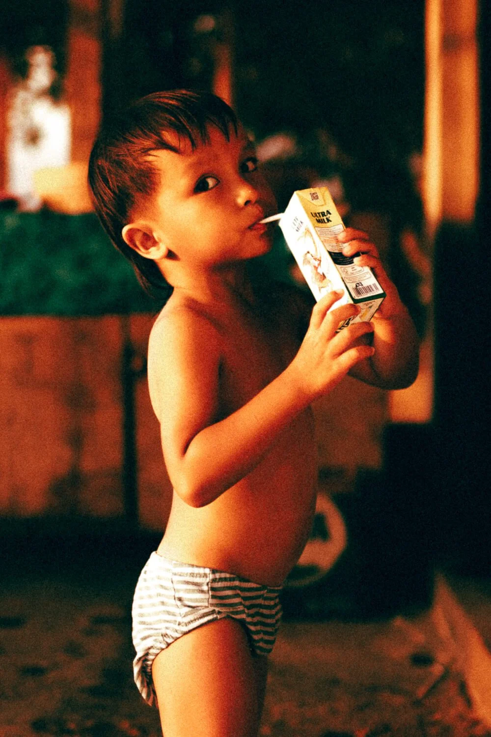 Un jeune garçon pieds nus et torse nu, portant un maillot de bain à rayures, boit du lait d'une boîte de lait en carton.