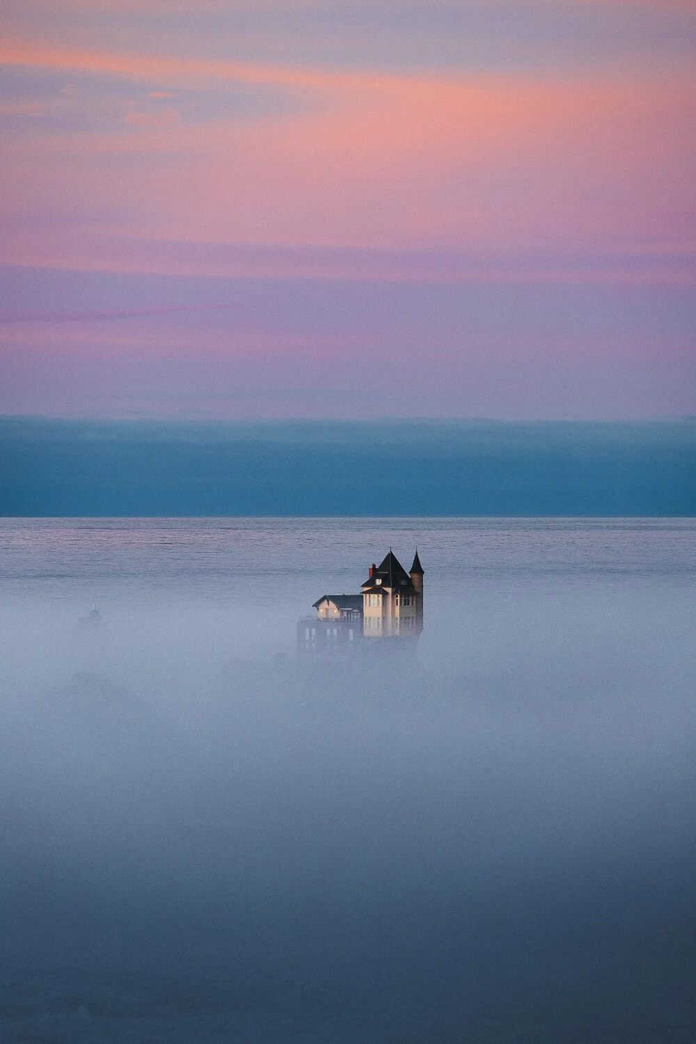 A house with a tower stands in the water, surrounded by fog, with a pastel pink and purple sky and distant mountains in the background.