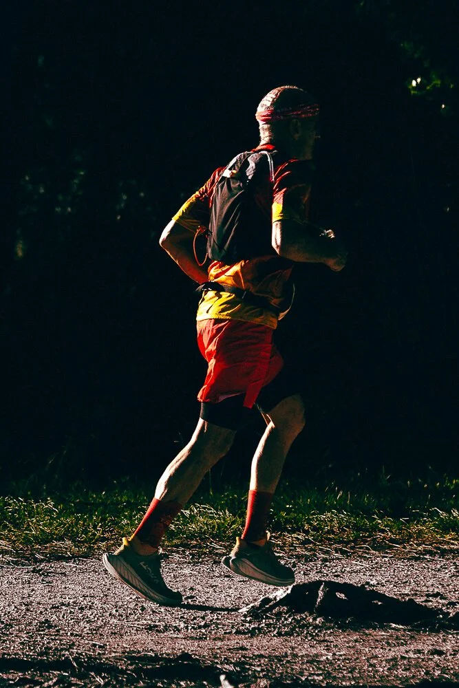 A man running on a trail at night, wearing a headband, red shorts, and a backpack.
