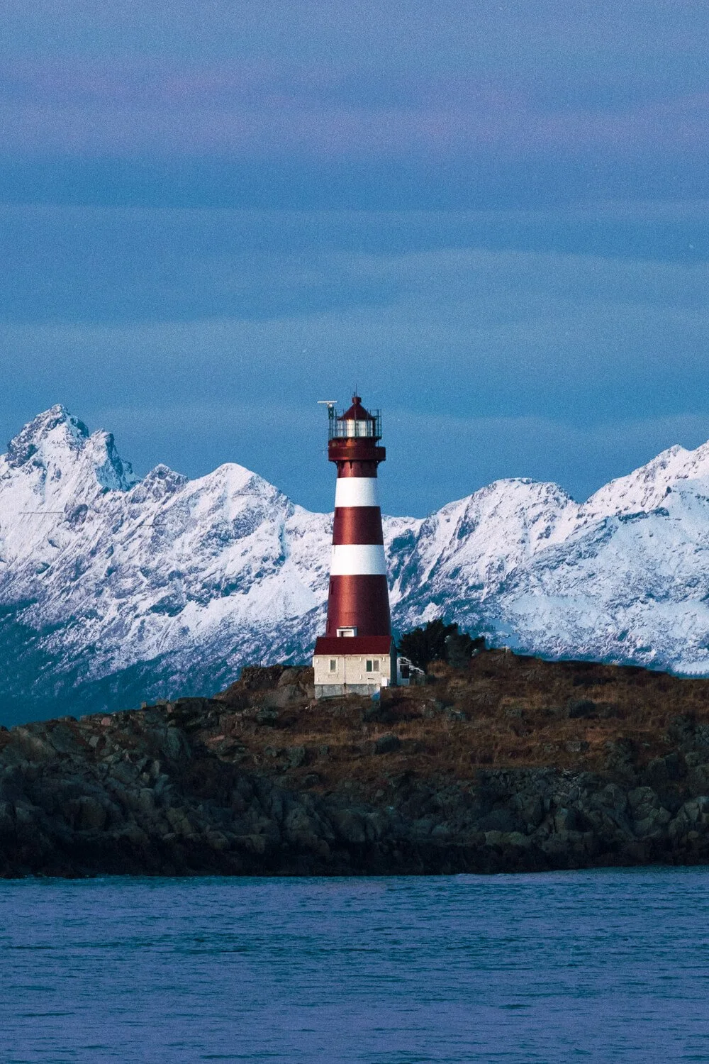 Un phare rouge et blanc situé sur une île rocheuse avec des montagnes enneigées en arrière-plan.