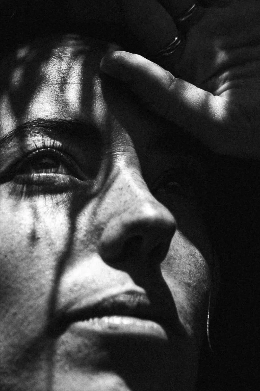 Close-up black and white photo of a woman with her eye closed, hand resting on her forehead, dramatic shadows highlighting her facial features.