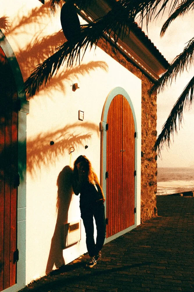 A woman standing against a white wall with a large wooden arched door, with shadows of palm leaves cast on the wall, during sunset near the beach.