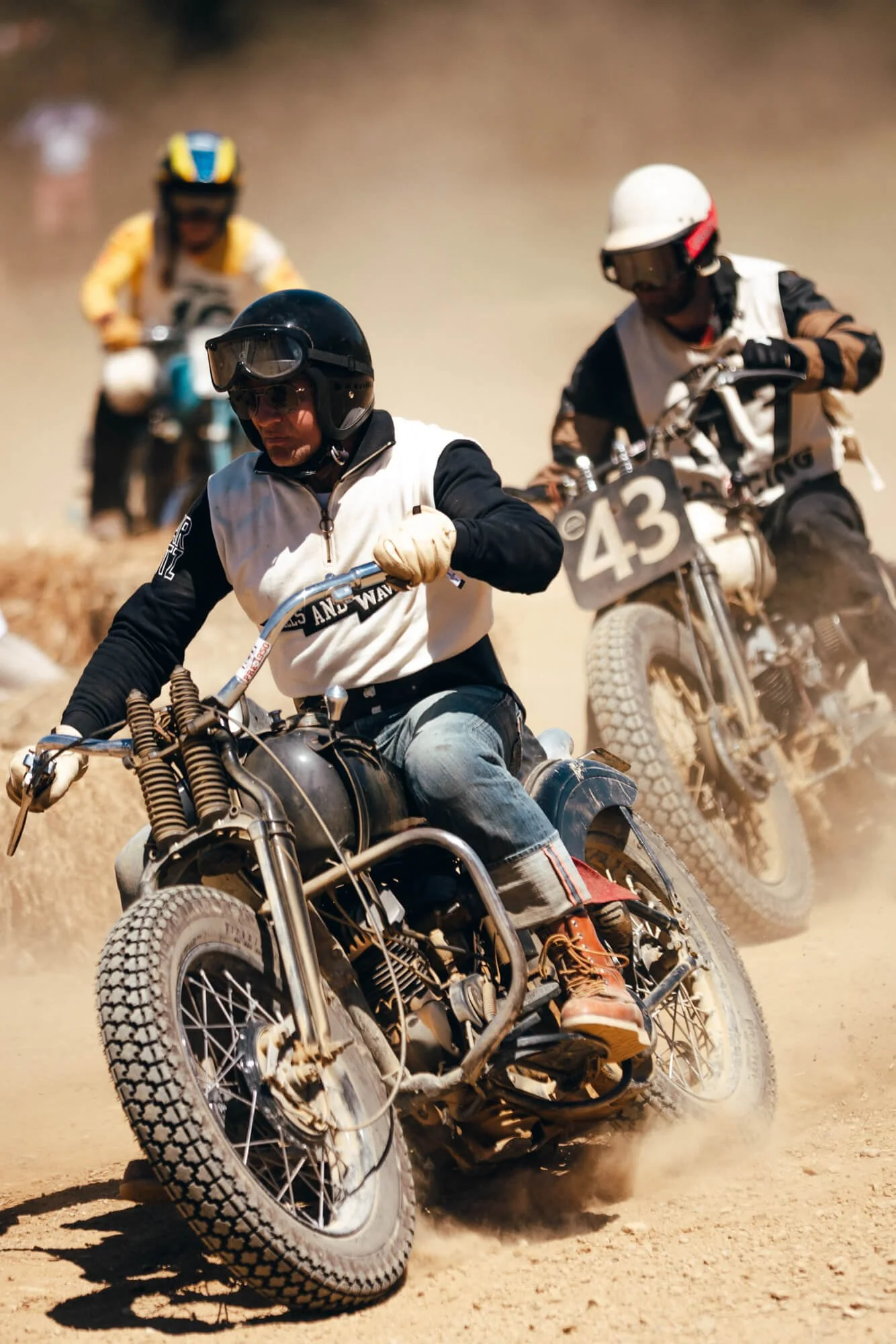Three motorcycle racers are racing on a dirt track, with two in the foreground and one in the background, kicking up dust as they speed around a bend.