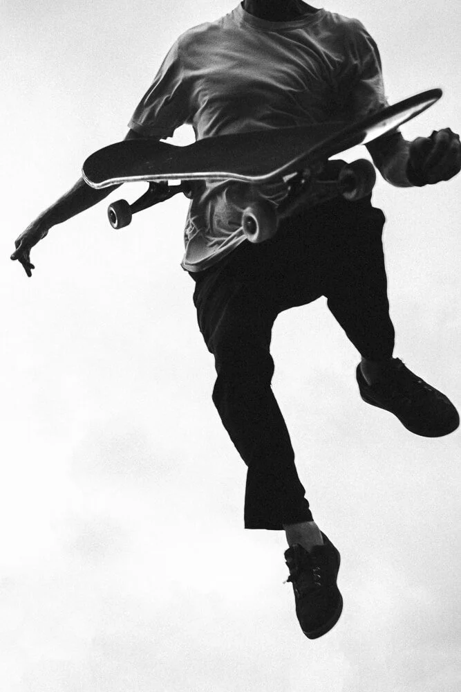 Person mid-jump holding a skateboard in their right hand, wearing a T-shirt and sneakers, against a plain background.