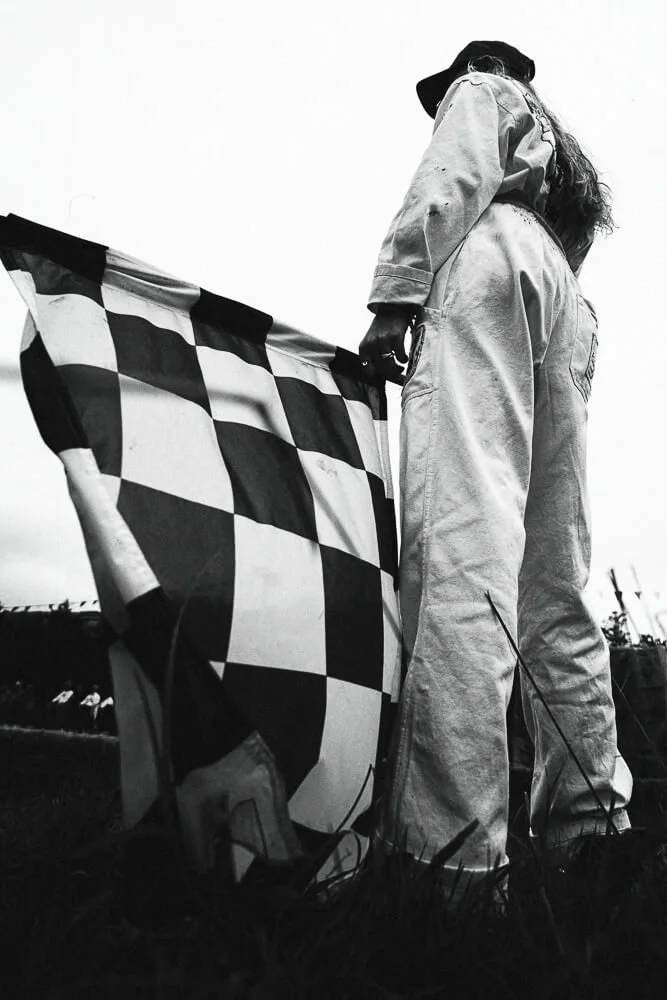 Person dressed in racing overalls holding a checkered racing flag outdoors.