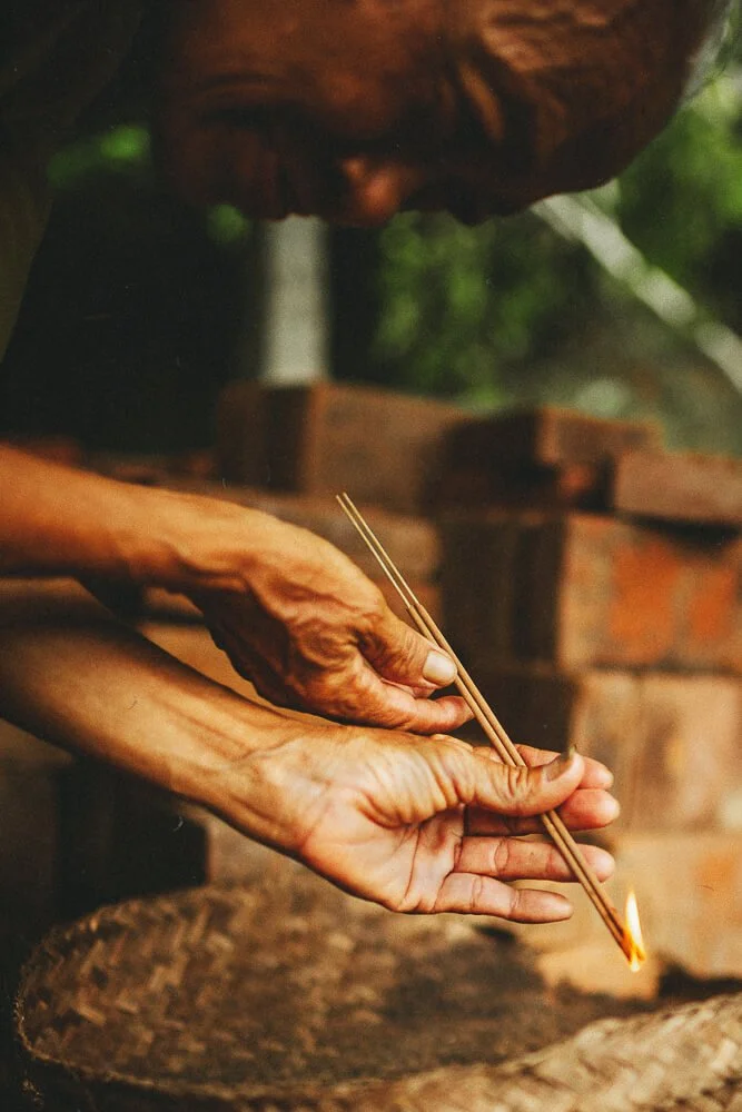 Two elderly hands hold incense sticks, with one stick burning at the tip, near a brick wall.