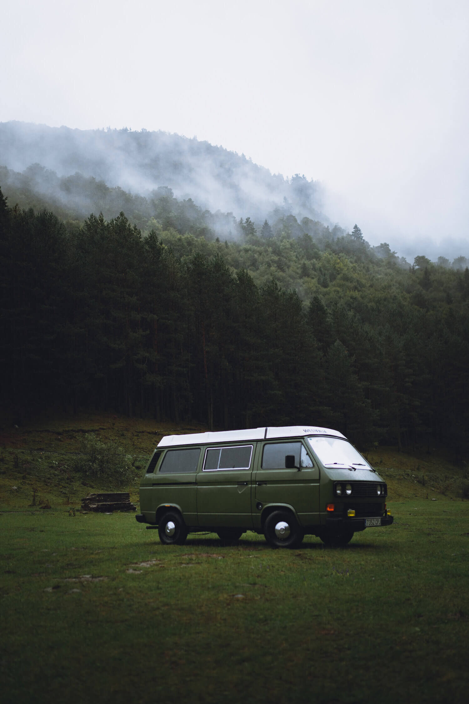 Un van vert stationné sur une pelouse devant une forêt brumeuse et des collines boisées.