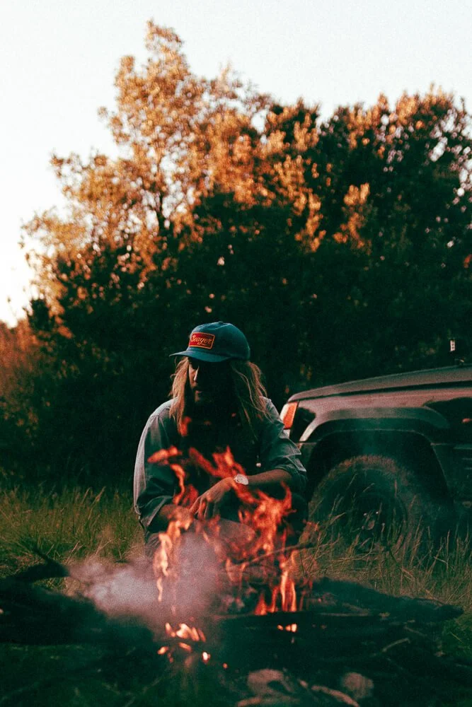Person with long hair wearing a baseball cap, crouching near a campfire outdoors during sunset with trees and a vehicle in the background.