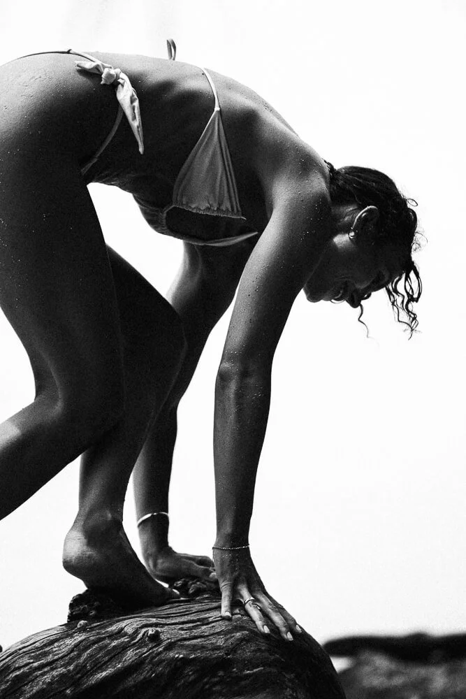 A woman in a swimsuit crouches on a large rock, reaching forward with one hand and balancing on her toes, with water droplets on her skin, against a plain background.