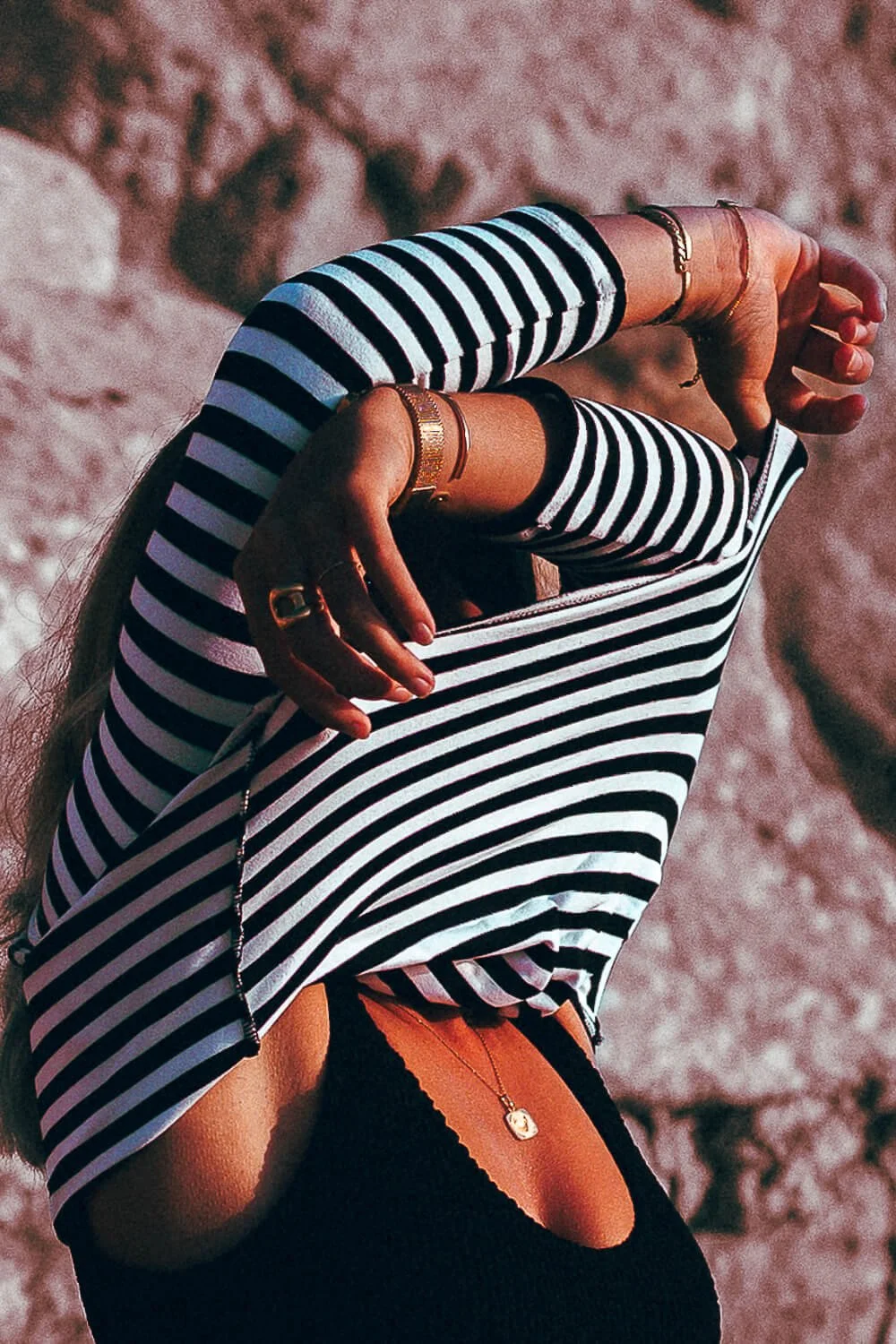 Person wearing a black and white striped long-sleeve shirt with their hands covering their face and head, standing outdoors in rocky terrain.
