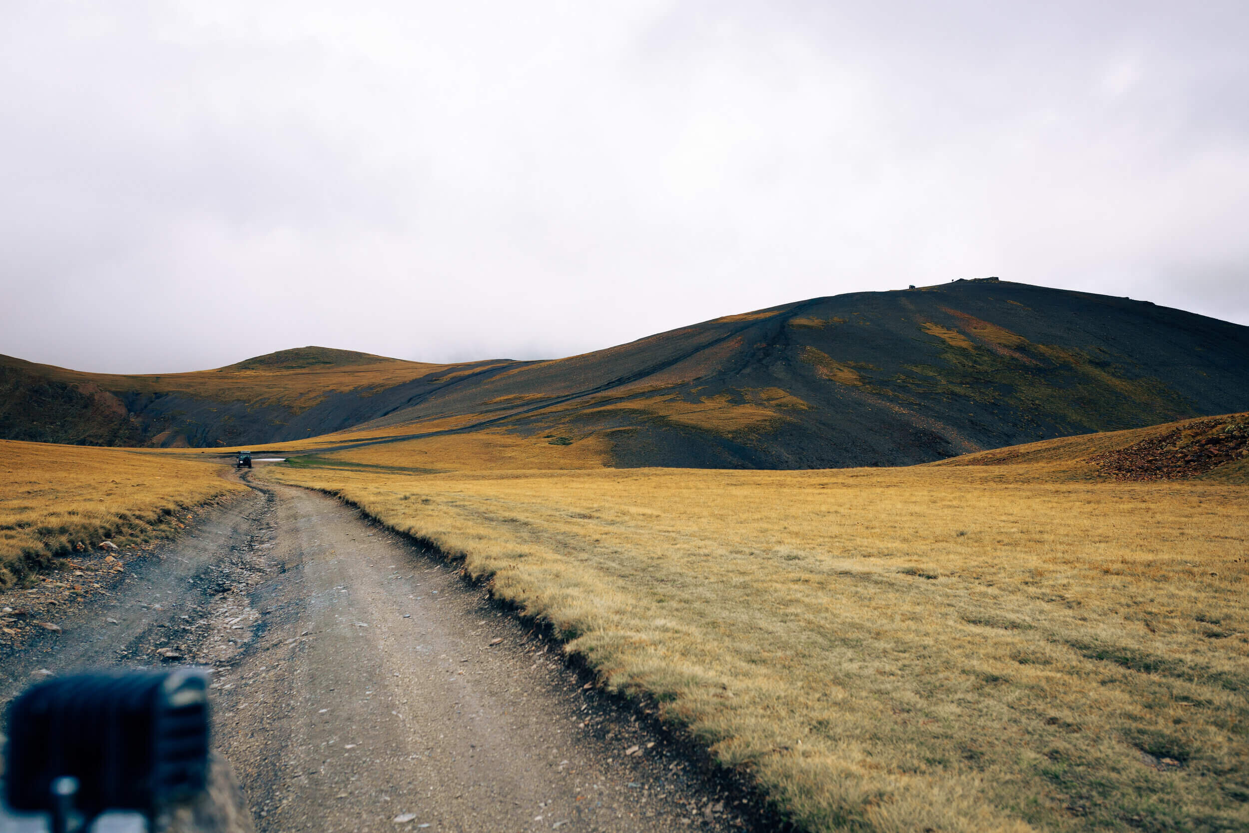Route de terre en montagne avec collines herbeuses et ciel nuageux.