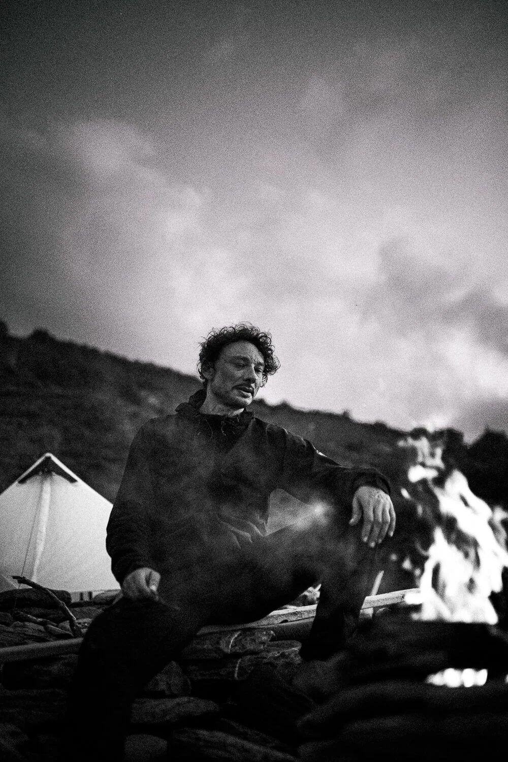A man with curly hair sitting by a campfire outdoors with tents and a hill in the background, captured in black and white.