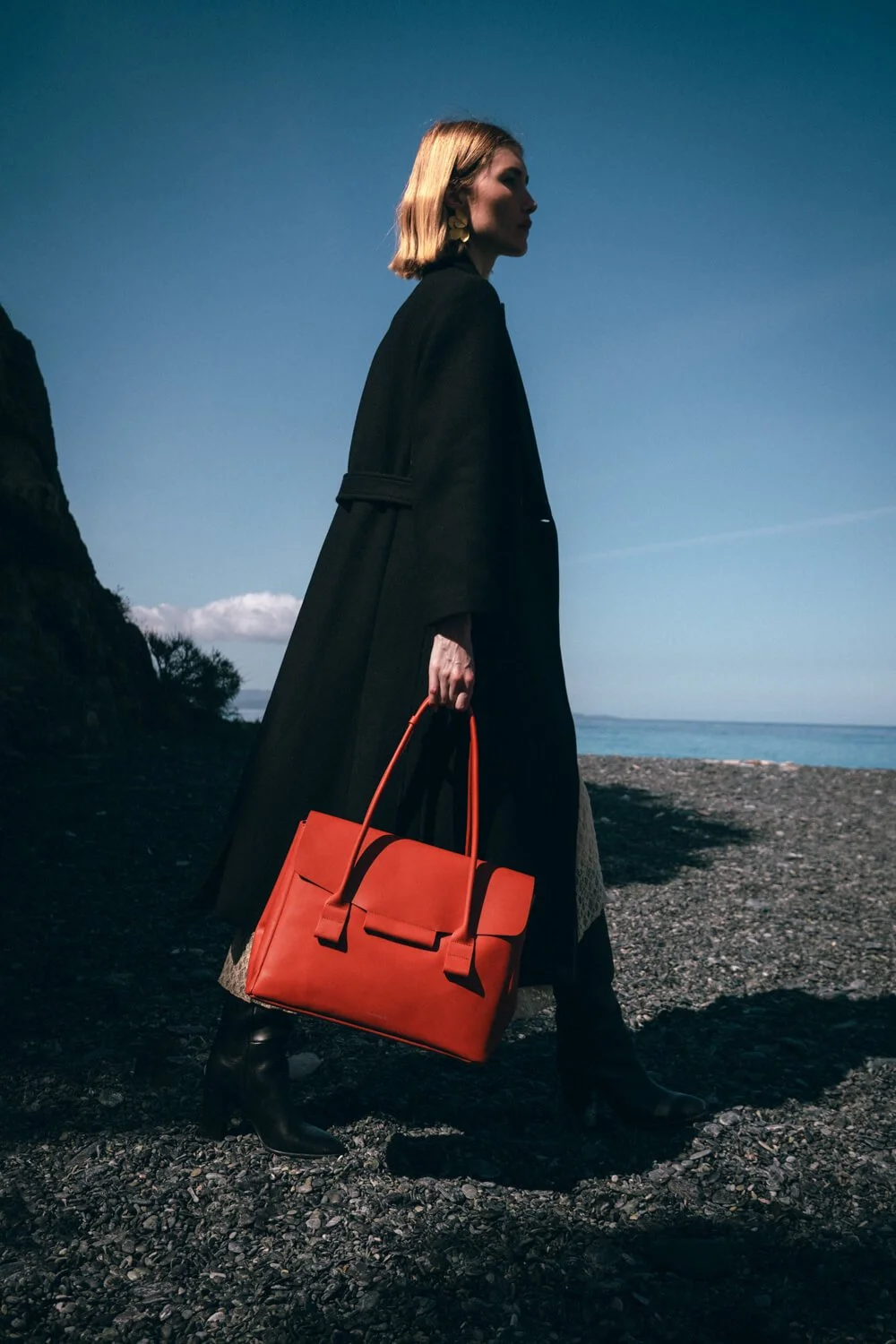 A woman in a black coat standing on a rocky beach, holding a red handbag, with the ocean and sky in the background.