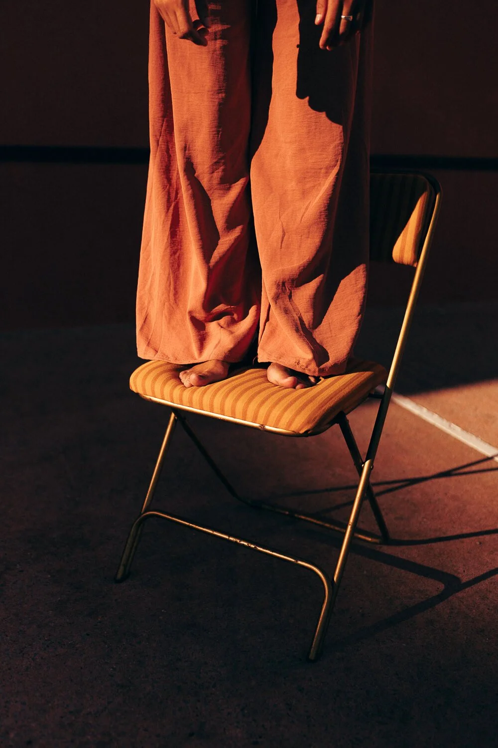 Person standing barefoot on a chair with fabric pants, with part of their hands visible, cast in warm, reddish lighting.