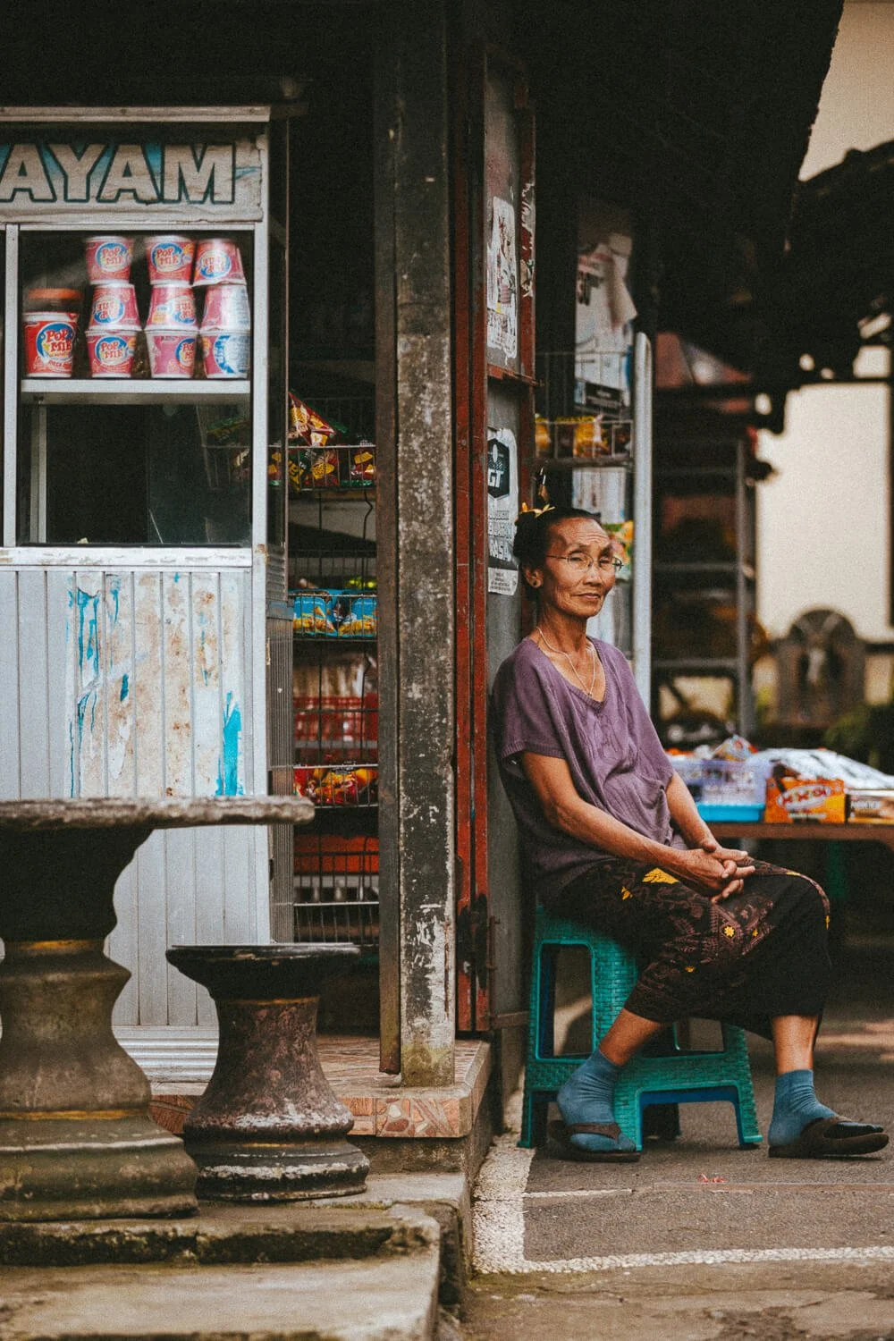 Une femme âgée assise sur une petite chaise en plastique devant un petit magasin d'épicerie avec des produits en arrière-plan, à l'extérieur, dans une atmosphère chaleureuse et rustique.
