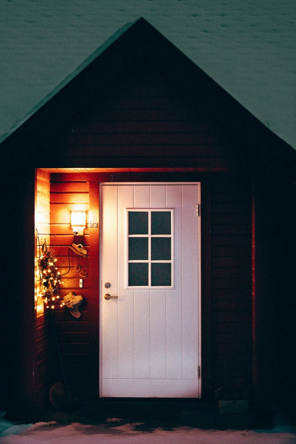 Porte de maison en bois blanc avec fenêtre, éclairage chaud, décoration de Noël avec petites lumières et sapin, maison en bois sombre, devant du sol enneigé.