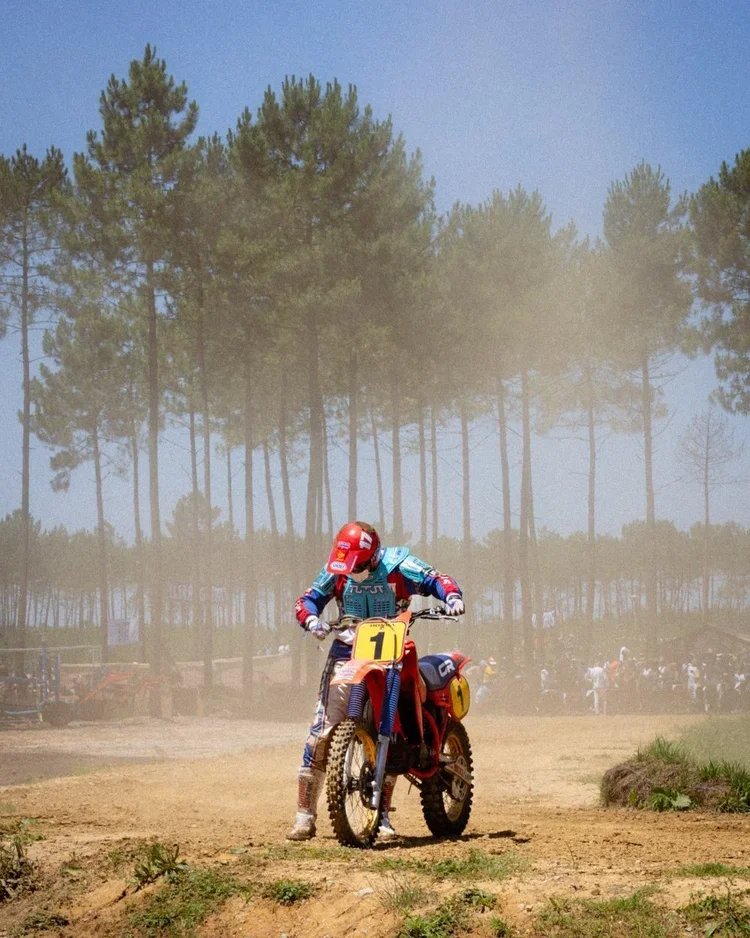 A motocross rider wearing a red helmet and blue gear sitting on a dirt bike with the number 1 on the front, on a dirt track with trees in the background.