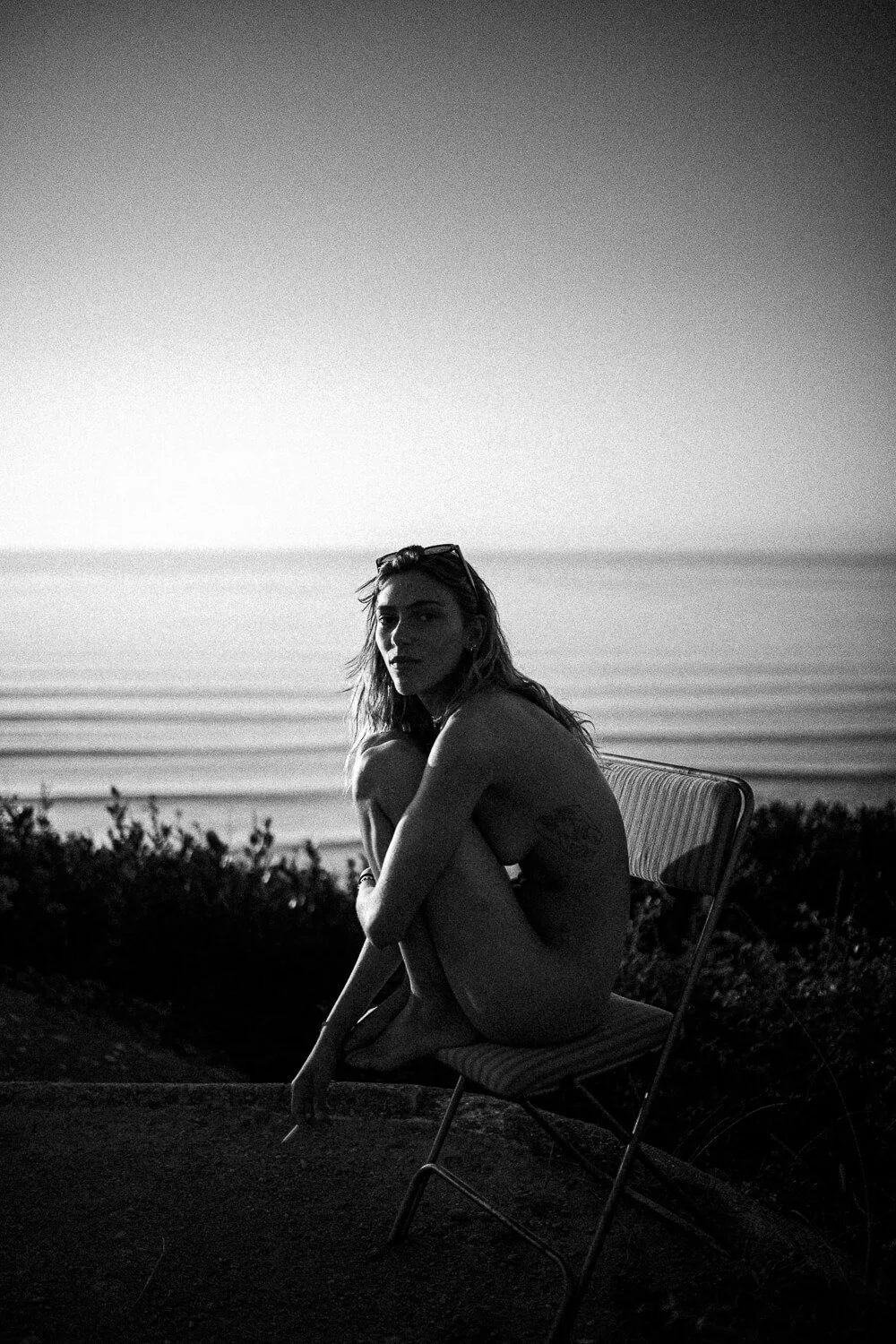Black and white photograph of a woman sitting nude on a chair near the beach, with the ocean in the background and the sky above.