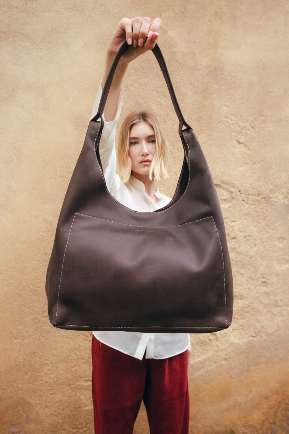 A young woman with blonde hair holding up a large dark brown leather tote bag in front of a beige textured wall.