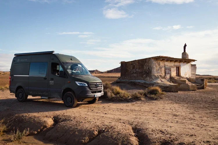 Un van Mercedes-Benz noir stationné à côté d'une vieille maison en pierre dans un paysage désertique avec un ciel bleu clair.